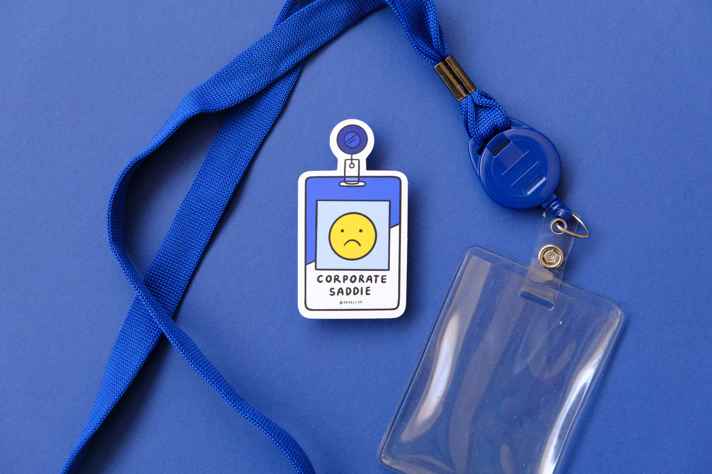 A JaneLi.Co sticker of a corporate badge that says "Corporate Saddie" with a blue lanyard and clear badge sleeve over a blue background.