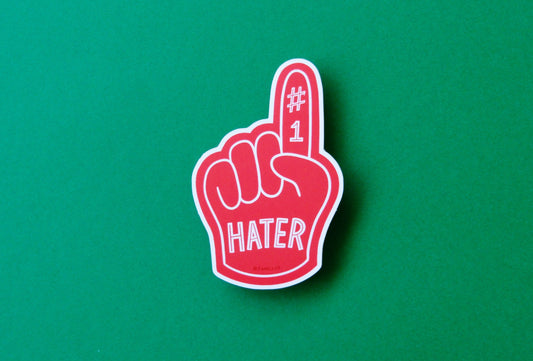A JaneLi.Co sticker of a red foam finger that says "#1 Hater" over a green background.
