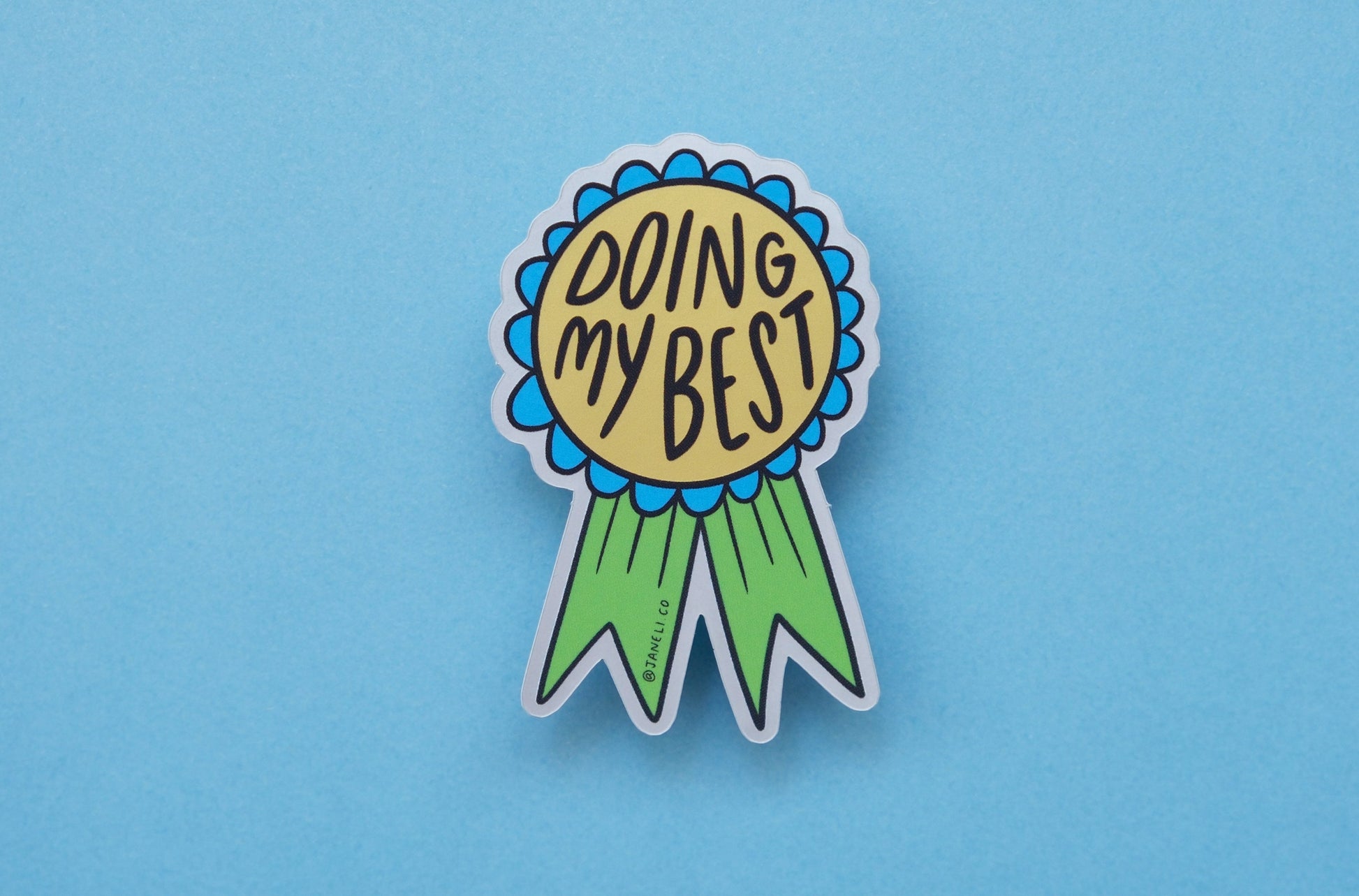 A JaneLi.Co sticker of a blue and green award ribbon that says "Doing My Best" over a blue background.