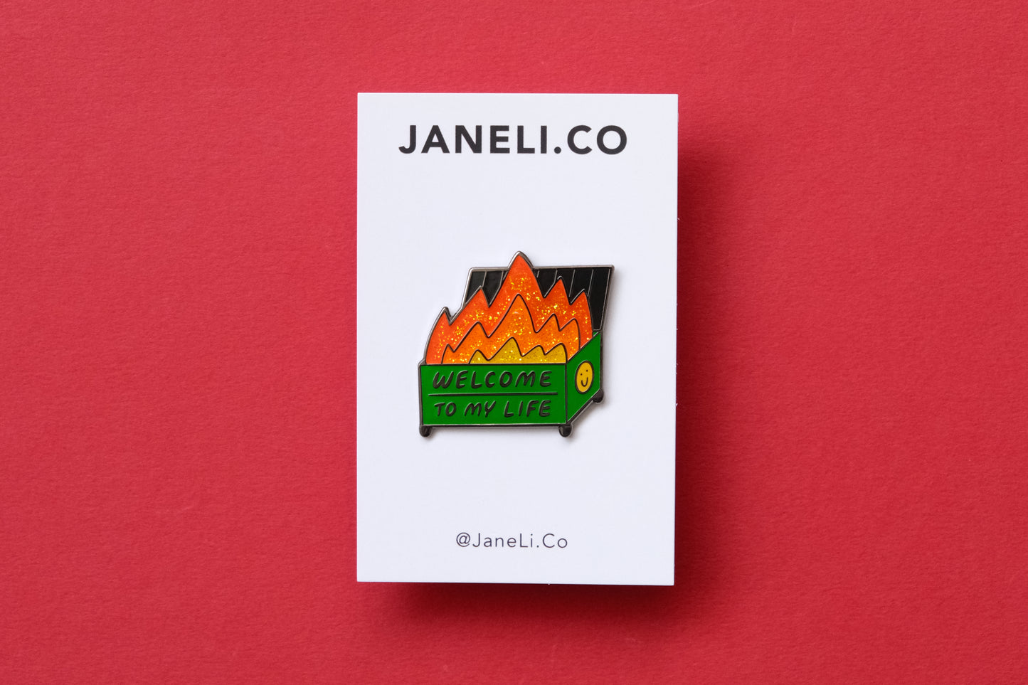 An enamel pin showing a dumpster with glittery flames that says "Welcome to my life" on a white JaneLi.Co backing card over a red background.