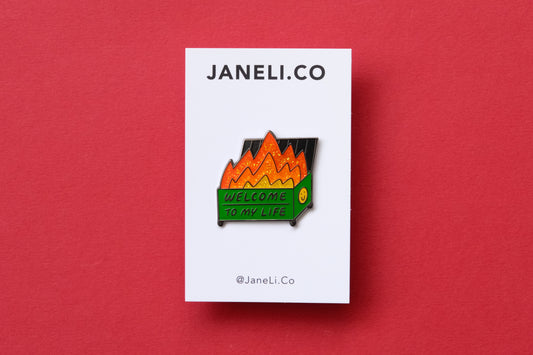 An enamel pin showing a dumpster with glittery flames that says "Welcome to my life" on a white JaneLi.Co backing card over a red background.