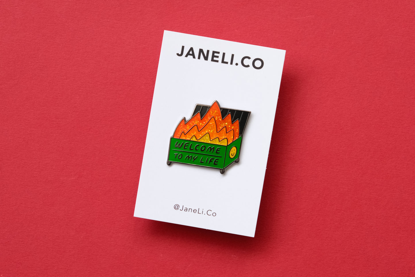 An enamel pin showing a dumpster with glittery flames that says "Welcome to my life" on a white JaneLi.Co backing card over a red background.