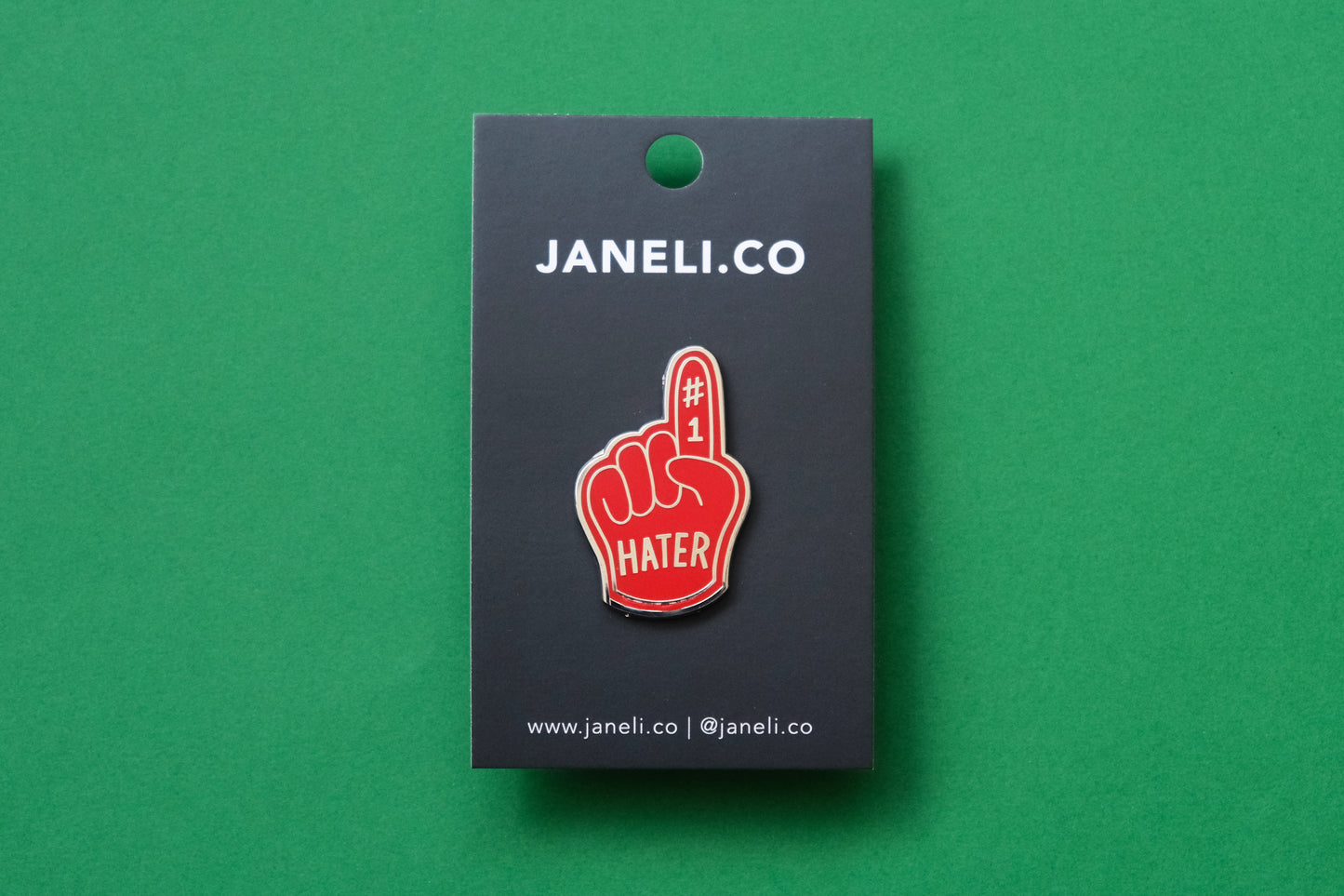 An enamel pin showing a red foam finger that says "#1 Hater" on a black JaneLi.Co backing card over a green background.