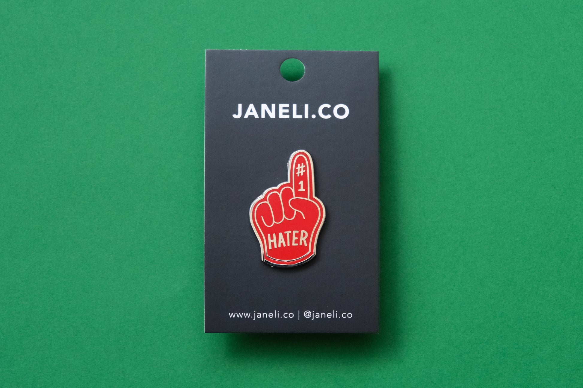 An enamel pin showing a red foam finger that says "#1 Hater" on a black JaneLi.Co backing card over a green background.