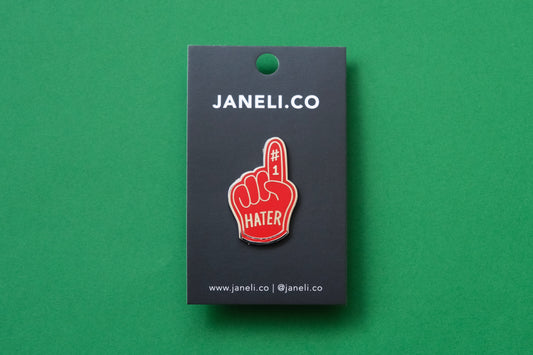 An enamel pin showing a red foam finger that says "#1 Hater" on a black JaneLi.Co backing card over a green background.