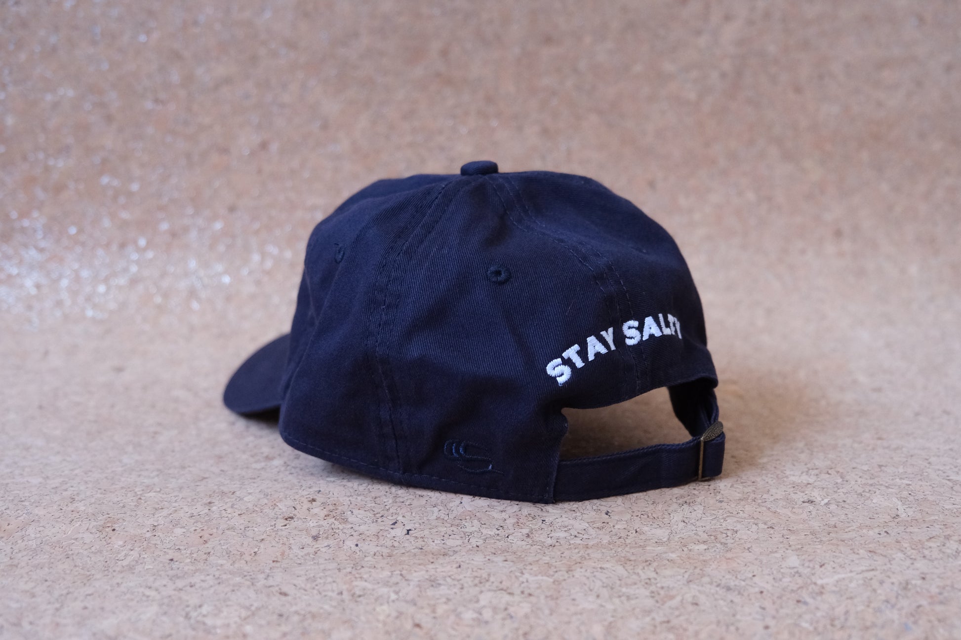 A photo of the back of a navy dad hat that says "Stay Salty" embroidered in white with an adjustable buckle strap.