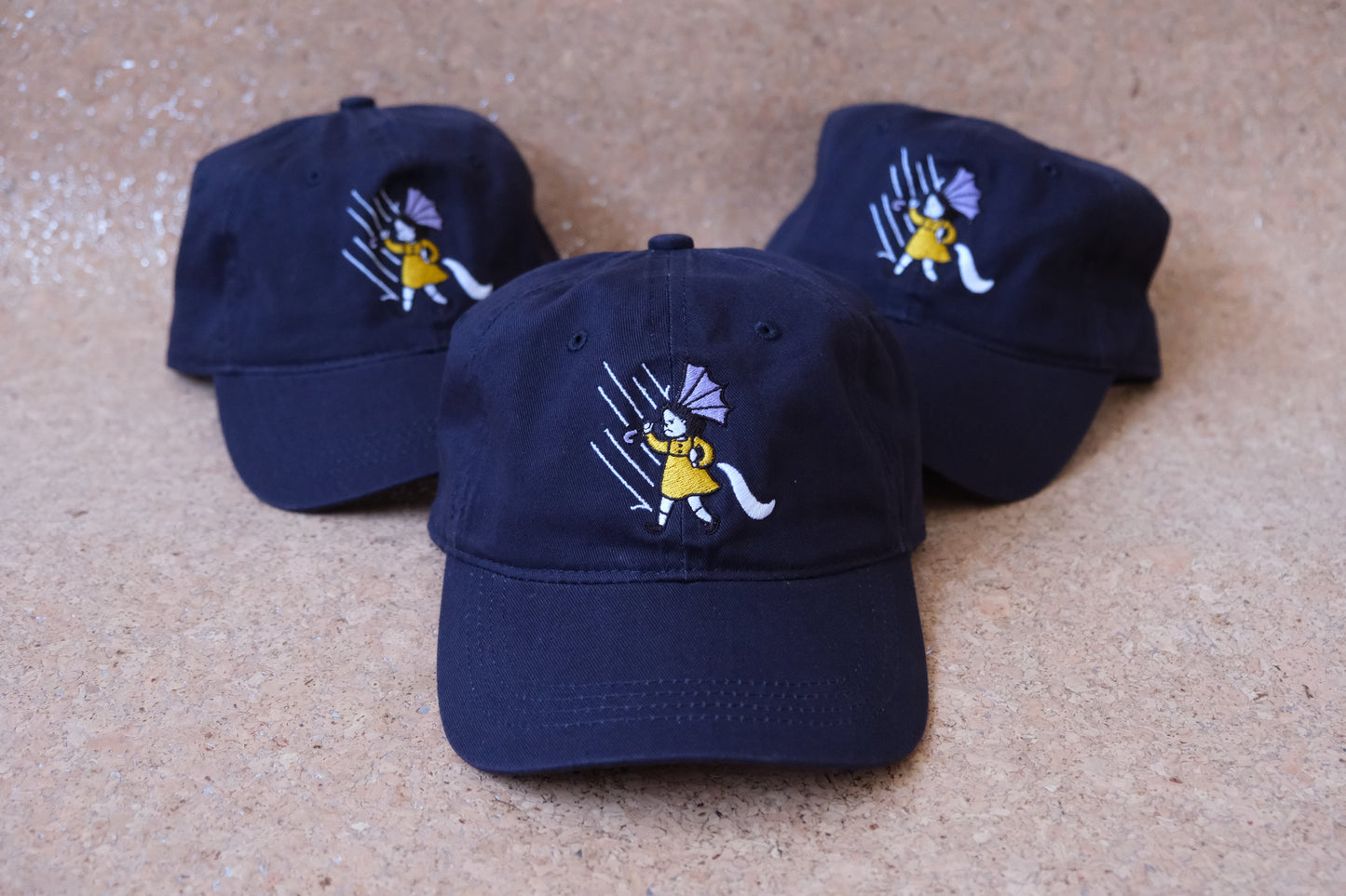 A photo of 3 navy dad hats with an embroidered grumpy salt girl over a tan background.