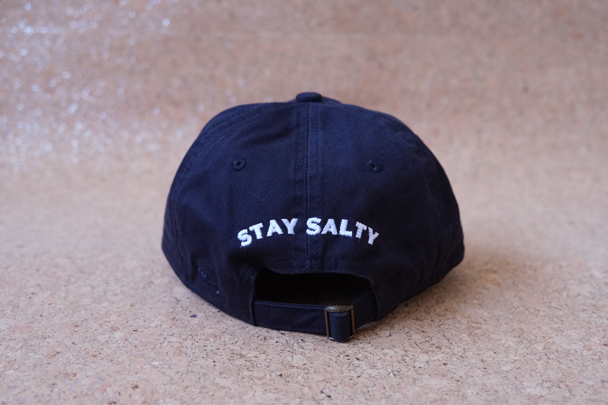 A photo of the back of a navy dad hat that says "Stay Salty" embroidered in white with an adjustable buckle strap.