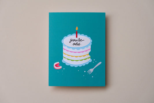 A photo of a teal greeting card with a birthday cake iced with "You're old." with a pair of dentures on a cream background.