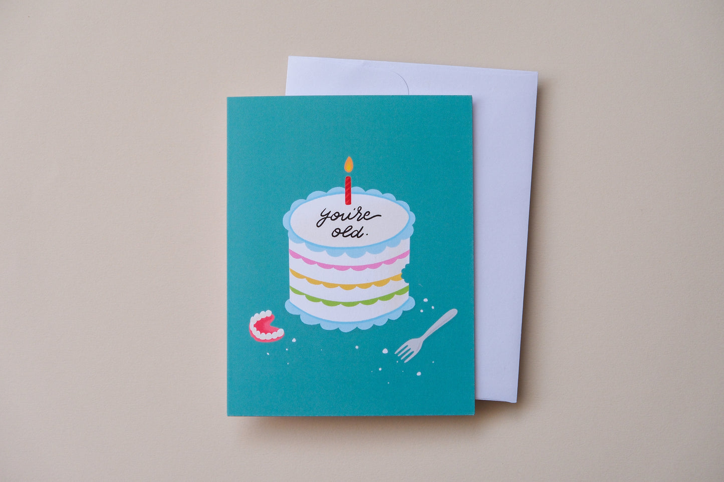 A photo of a teal greeting card with a birthday cake iced with "You're old." with a pair of dentures and a white envelope on a cream background.