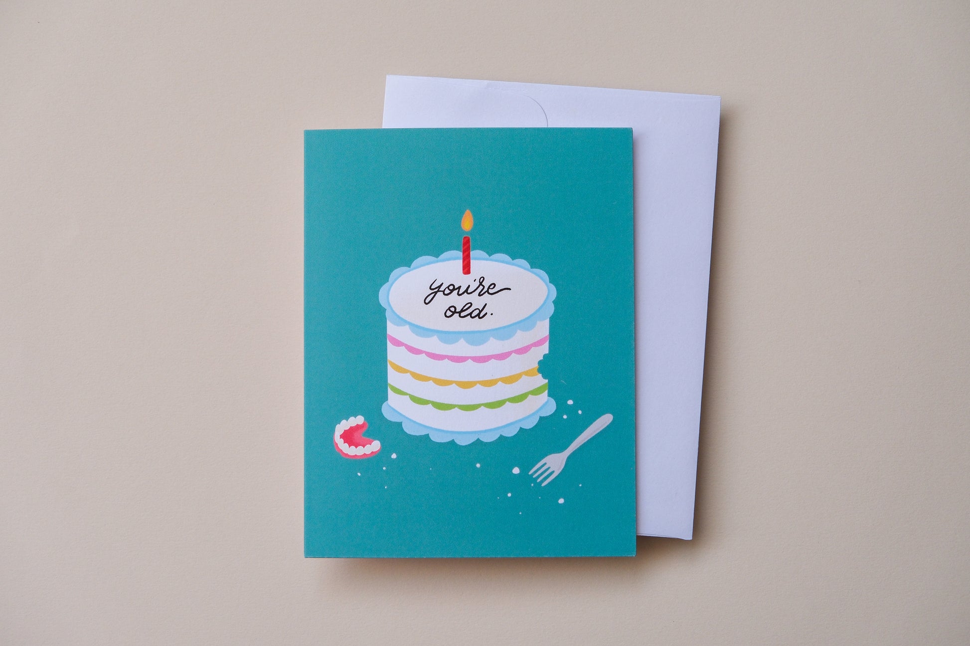 A photo of a teal greeting card with a birthday cake iced with "You're old." with a pair of dentures and a white envelope on a cream background.