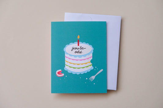 A photo of a teal greeting card with a birthday cake iced with "You're old." with a pair of dentures and a white envelope on a cream background.