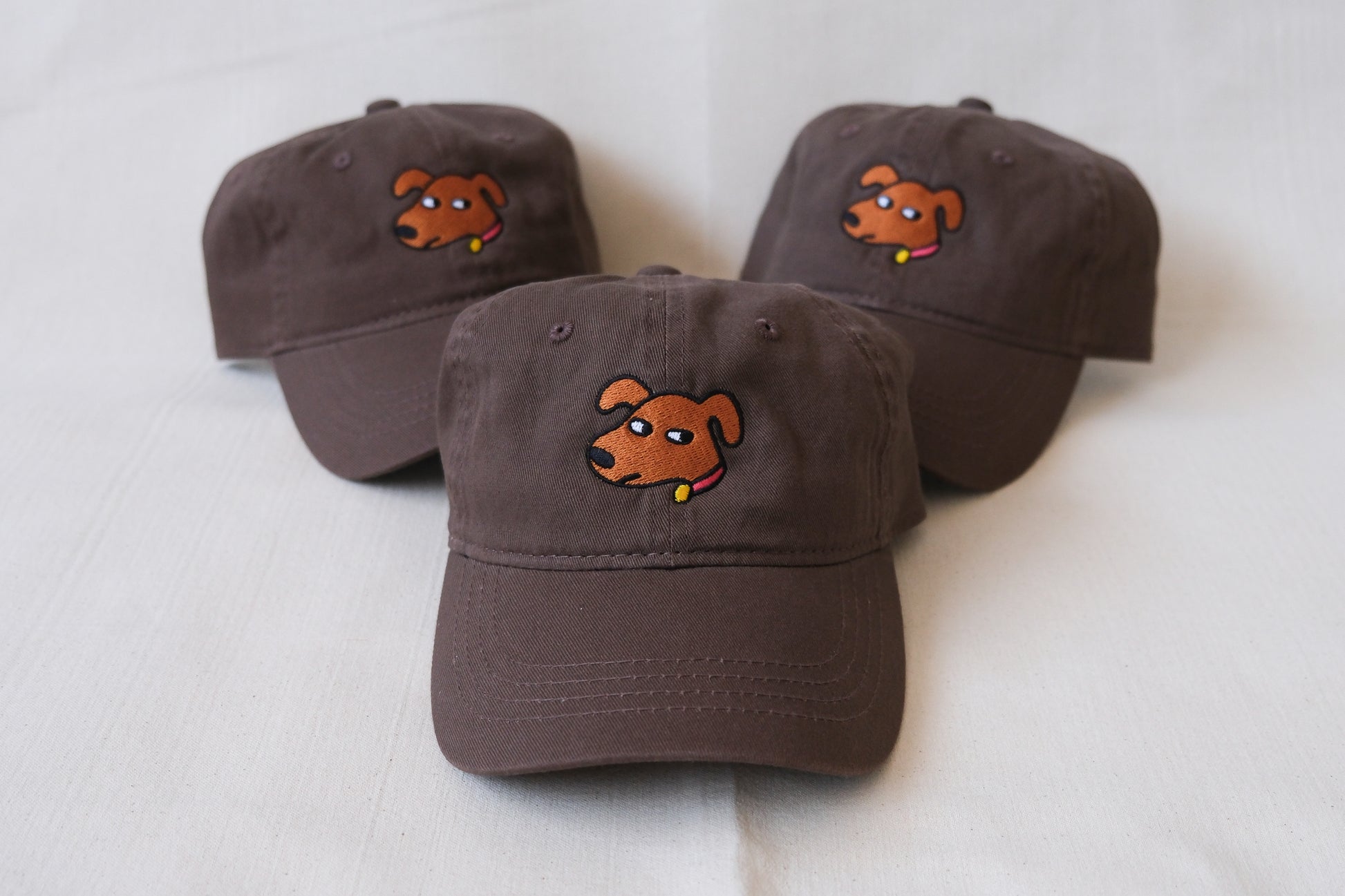 A photo of a 3 chocolate brown dad hats embroidered with dogs giving side eye over a cream background.