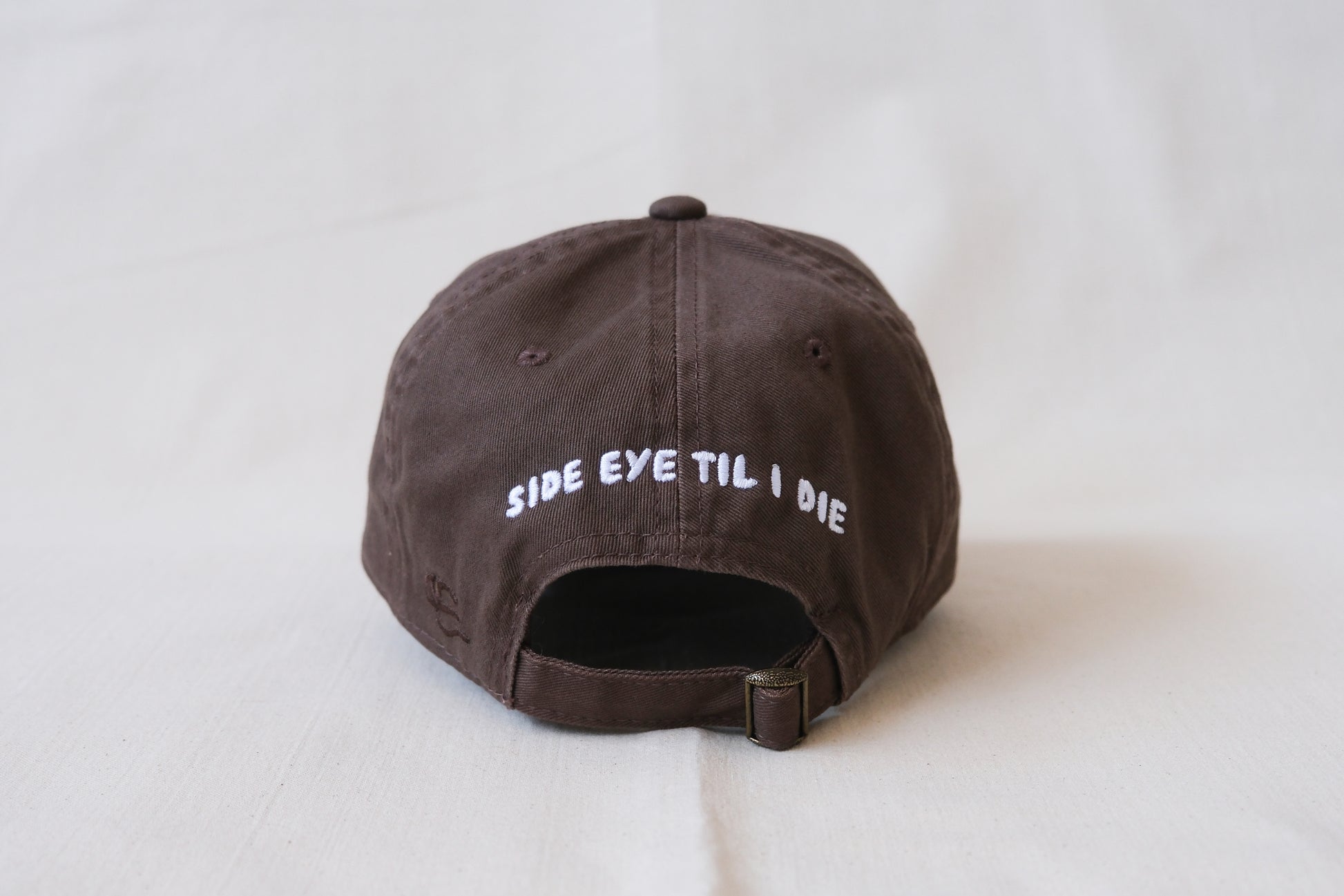 A photo of the back of a chocolate brown dad hat that says "Side Eye til I Die" embroidered in white with an adjustable buckle strap.
