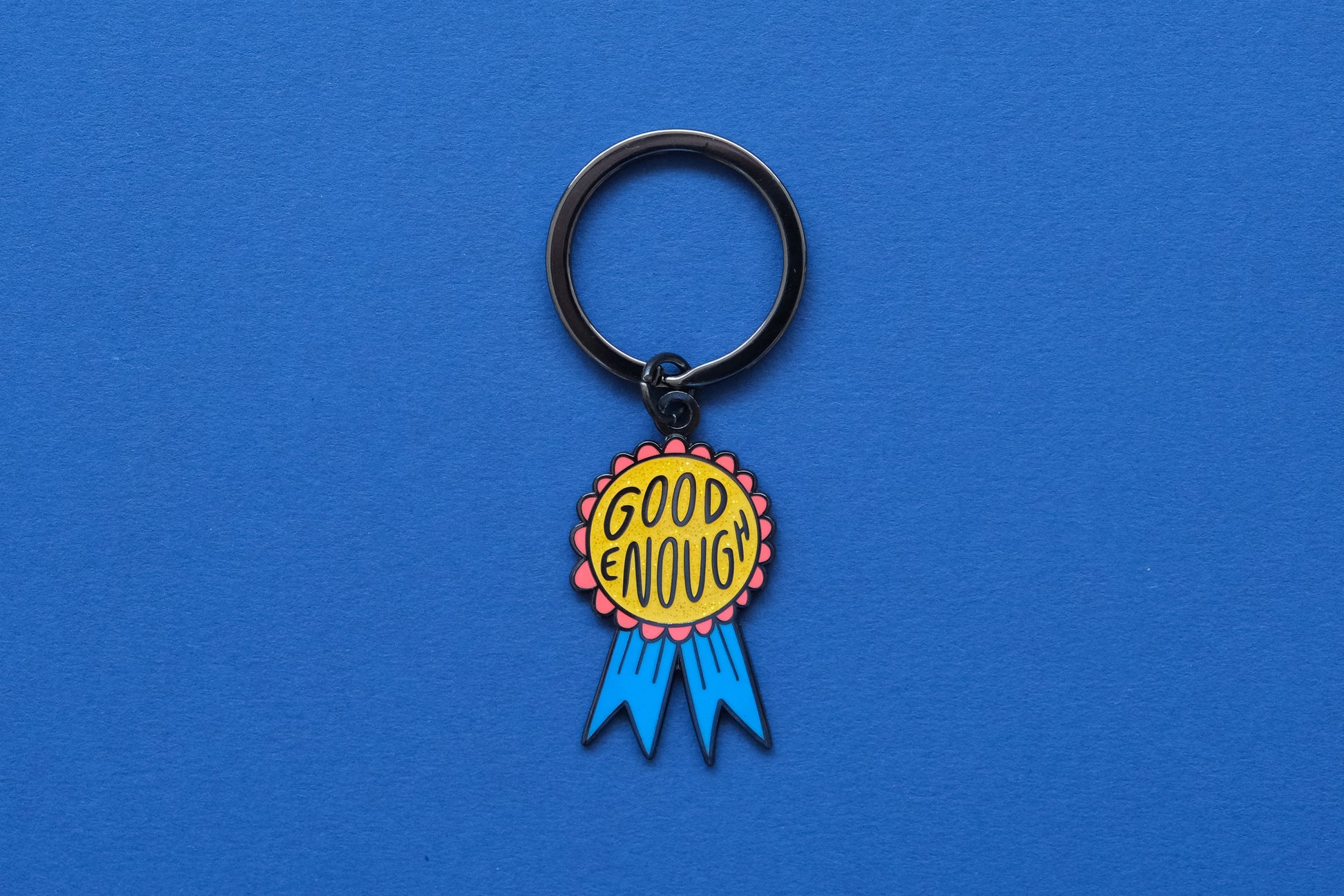 An enamel keychain showing a glittery red and blue award ribbon that says "Good Enough" over a blue background.