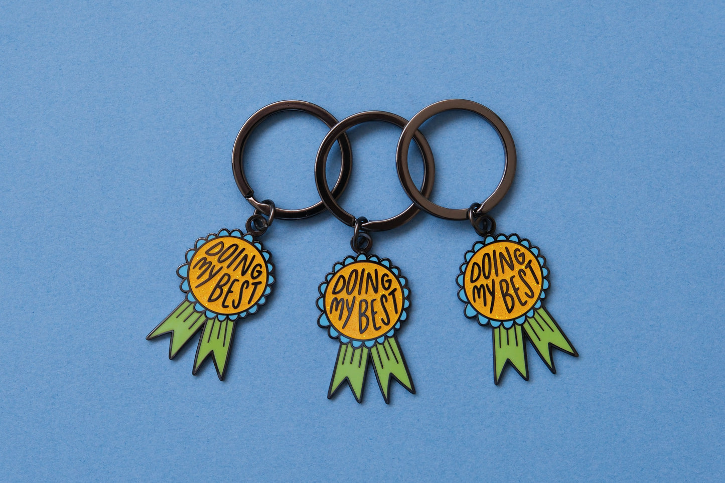 3 enamel keychains showing award ribbons that says "Doing My Best" over a blue background.