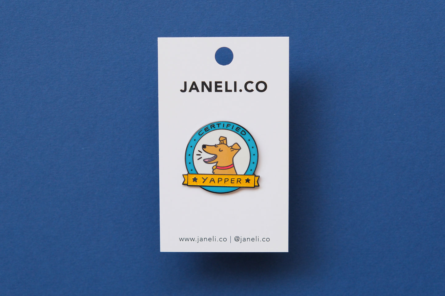 An enamel pin showing a barking dog that says "Certified Yapper" on a white JaneLi.Co backing card over a blue background.