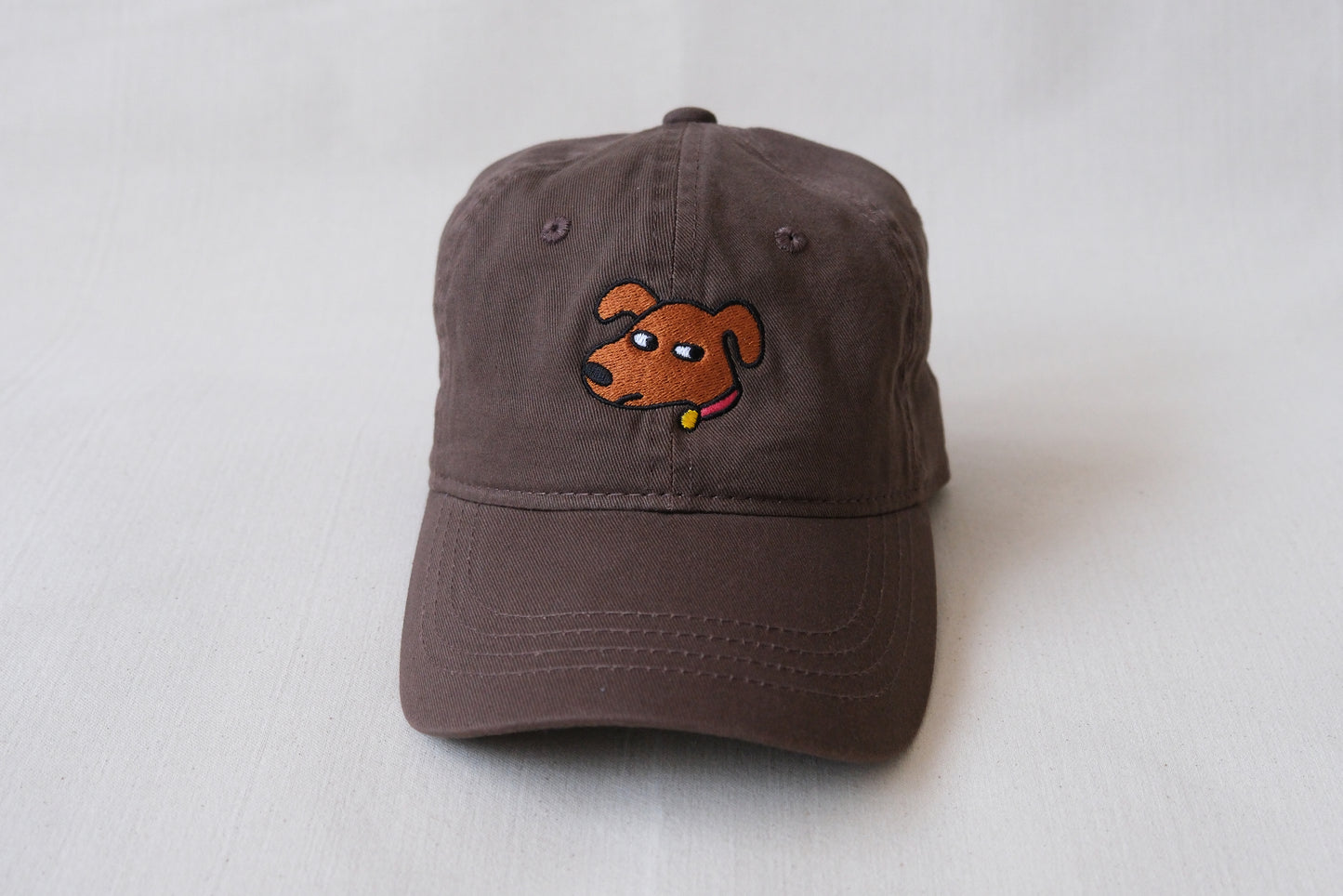 A photo of a chocolate brown dad hat with an embroidered dog giving side eye over a cream background.