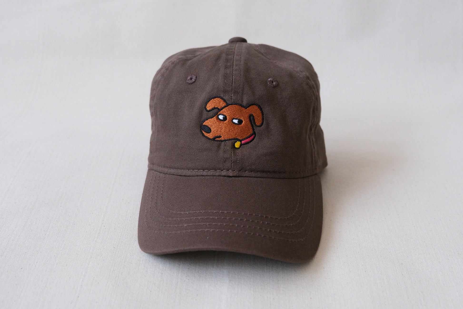 A photo of a chocolate brown dad hat with an embroidered dog giving side eye over a cream background.