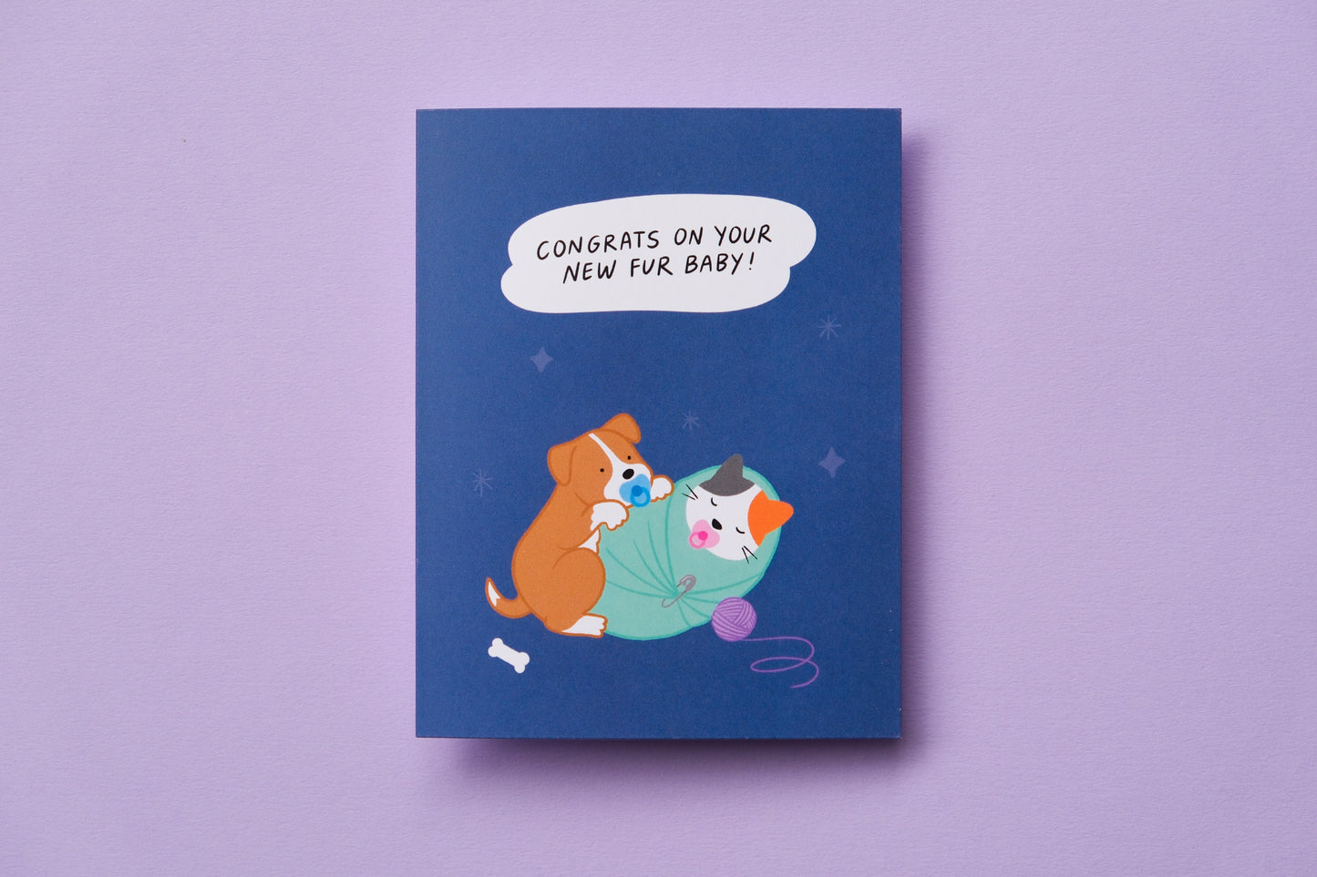 A photo of a greeting card that says "Congrats on your new fur baby!" with a puppy and a kitten swaddled and sucking on pacifiers on a lavender background.
