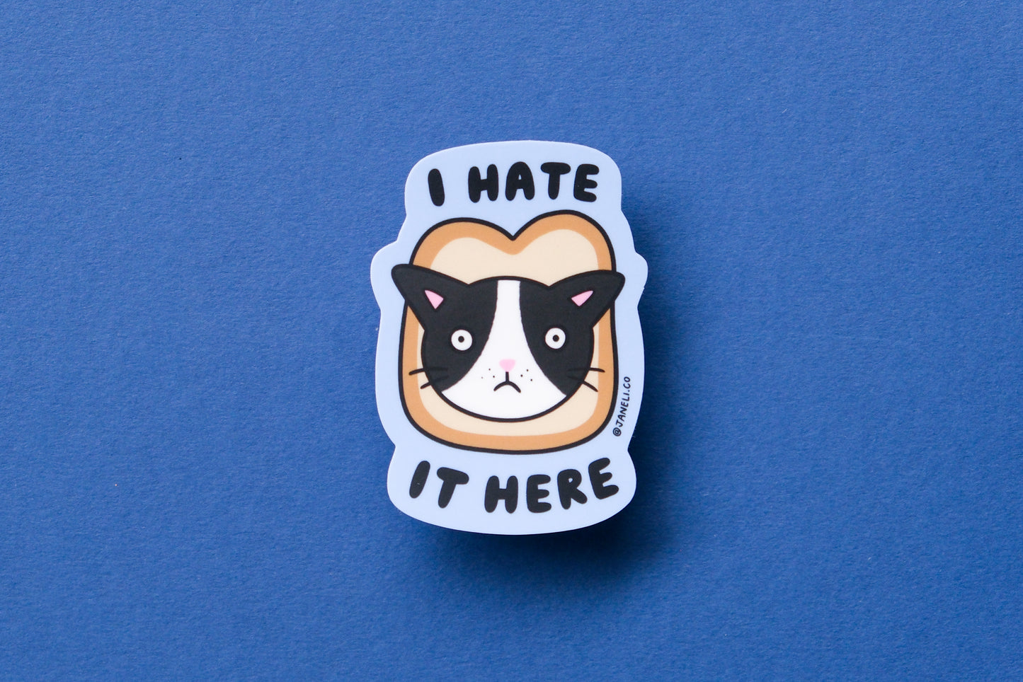 A JaneLi.Co sticker of a cat stuck in a slice of bread that says "I hate it here" over a blue background.