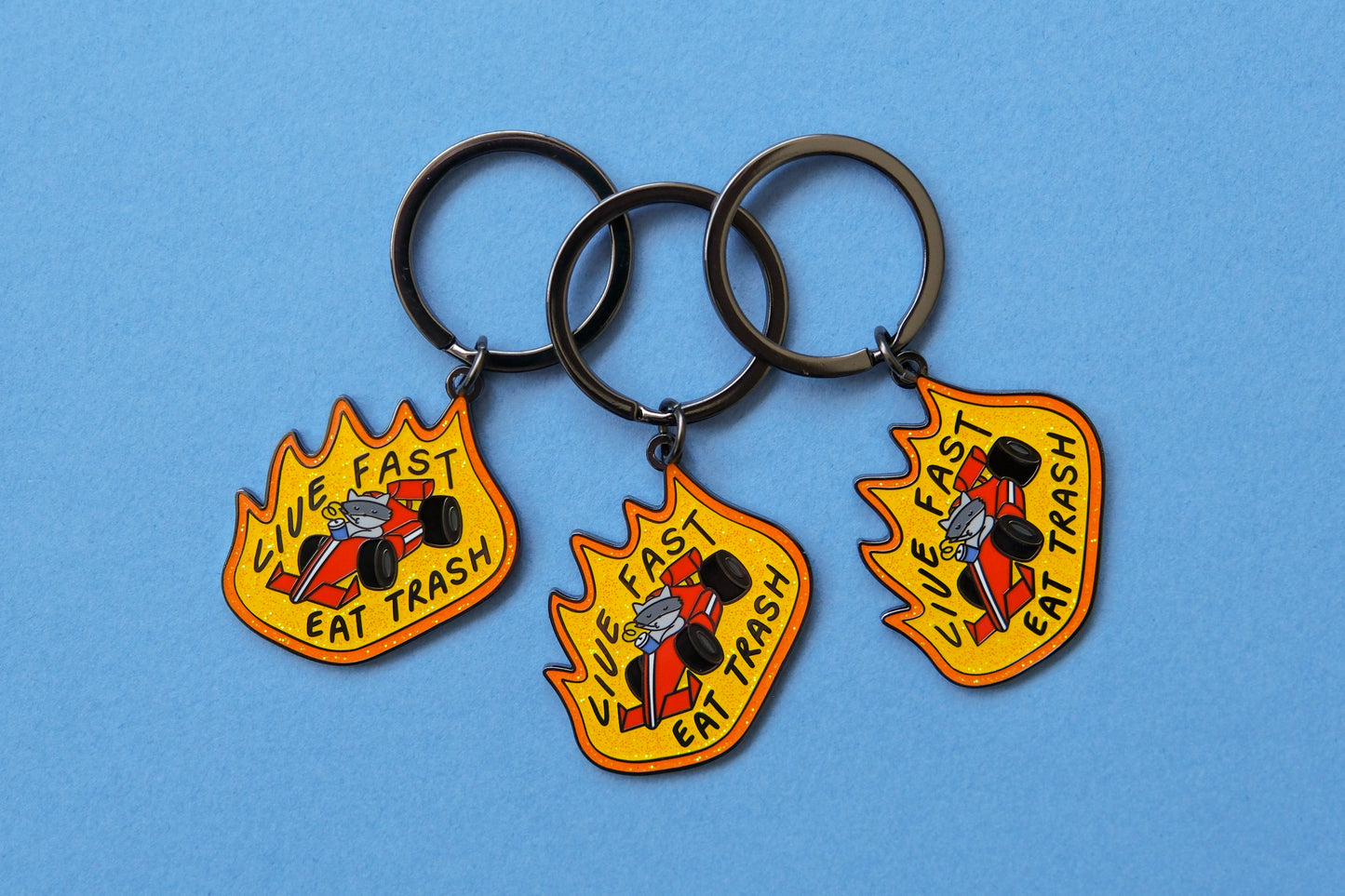 3 enamel keychains of raccoons driving a racecar while sipping a soda that says "Live Fast Eat Trash" over a light blue background.