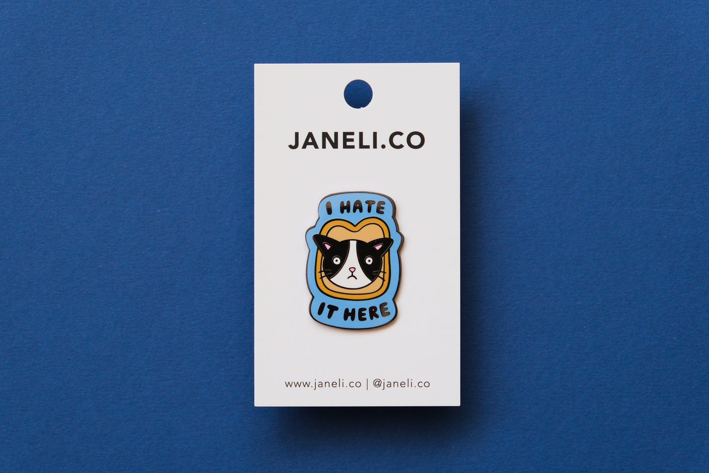 An enamel pin of a cat stuck in a piece of bread that says "I Hate It Here" on a white JaneLi.Co backing card over a blue background.