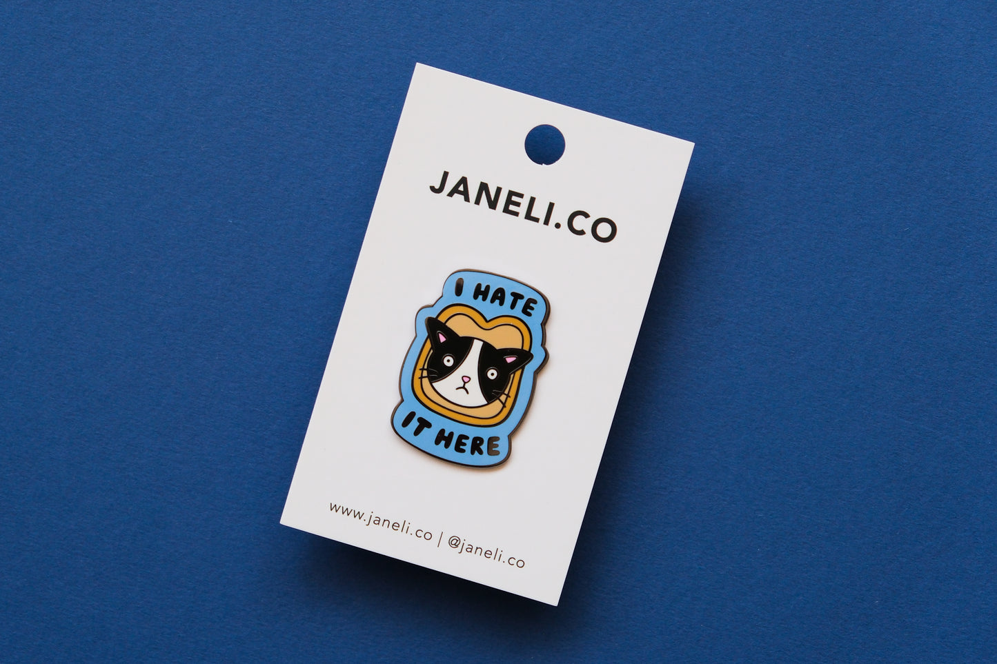 An enamel pin of a cat stuck in a piece of bread that says "I Hate It Here" on a white JaneLi.Co backing card over a blue background.