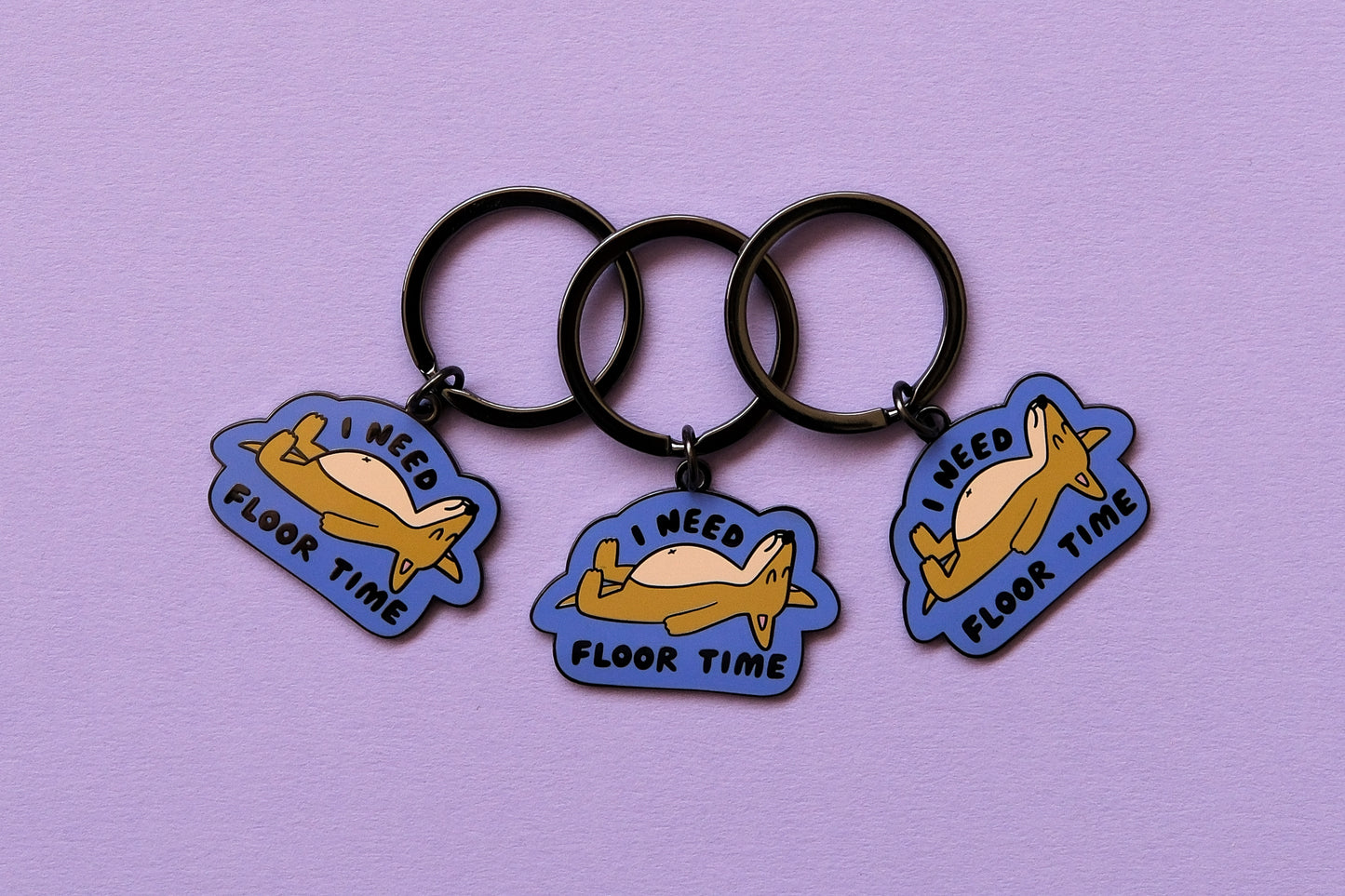 3 enamel keychains of dogs lying down that say "I Need Floor Time" over a lavender background.