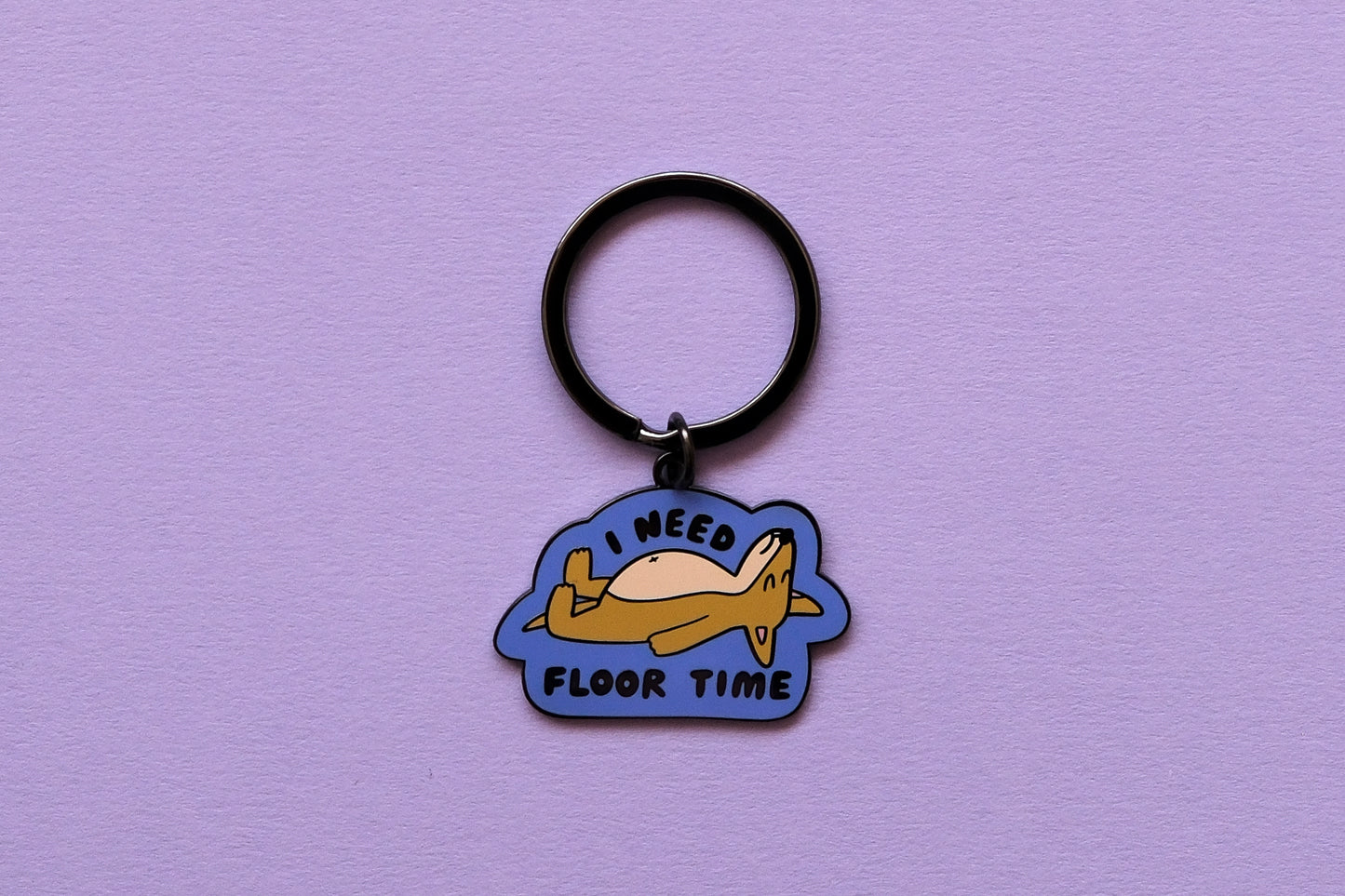 An enamel keychain of a dog lying down that says "I Need Floor Time" over a lavender background.
