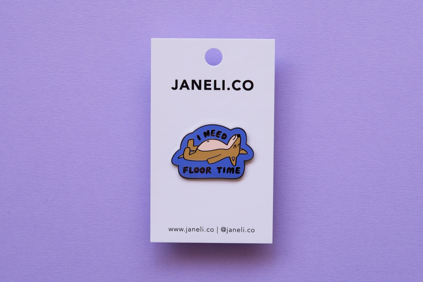 An enamel pin of a dog lying down that says "I Need Floor Time" on a white JaneLi.Co backing card over a lavender background.