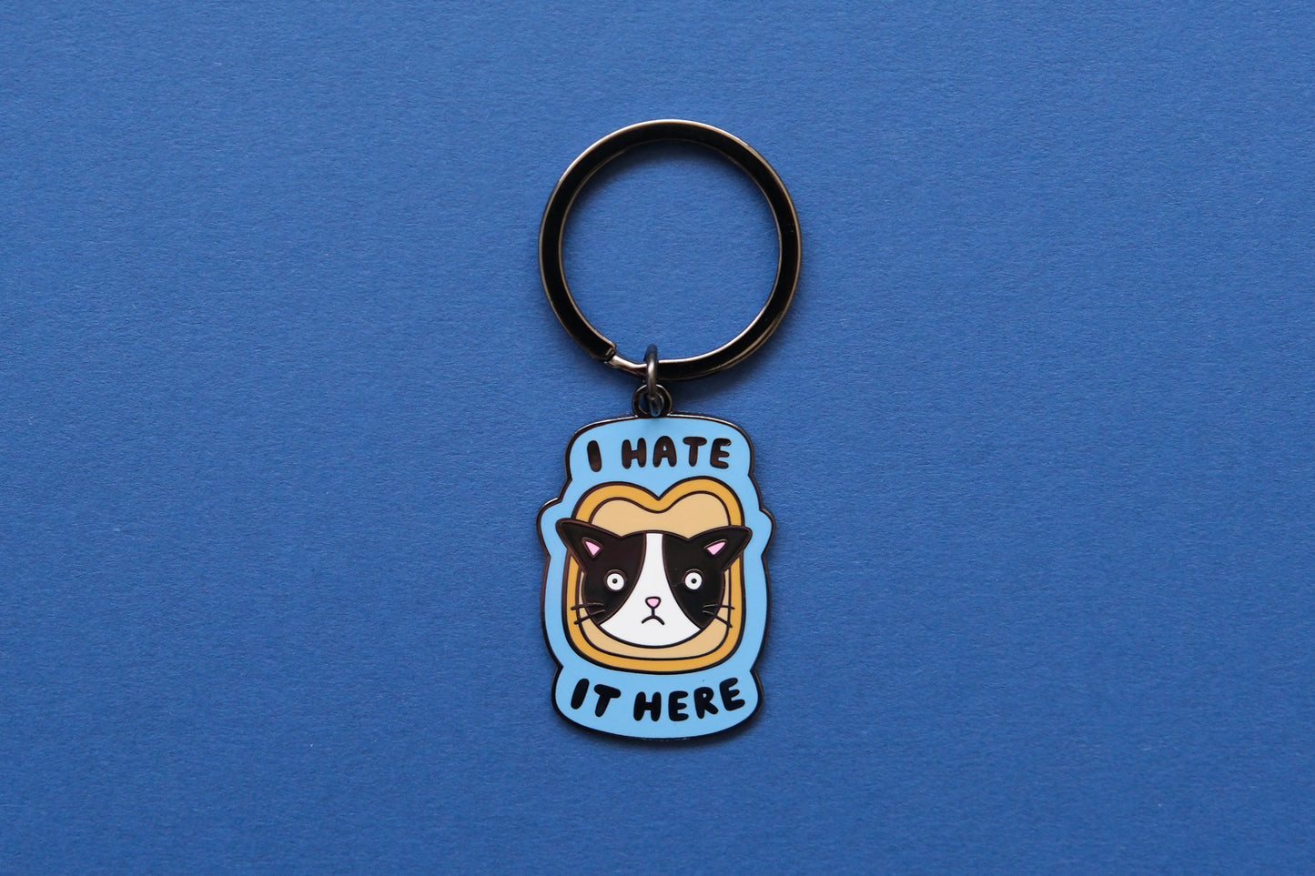 An enamel keychain of a cat stuck in a piece of bread that says "I Hate It Here" over a blue background.