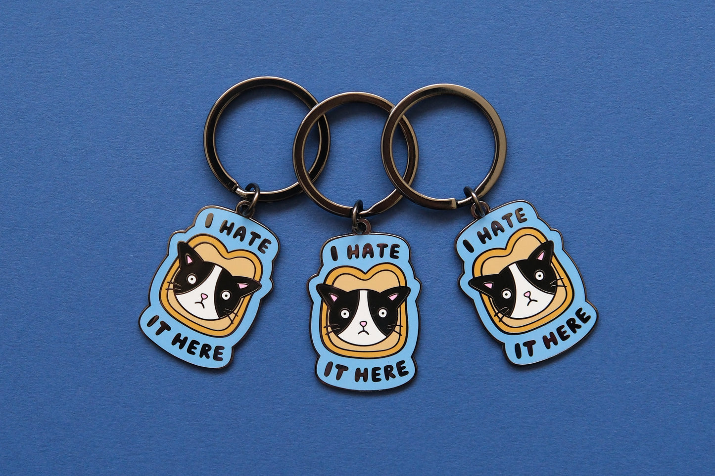 3 enamel keychains of cats stuck in a piece of bread that say "I Hate It Here" over a blue background.