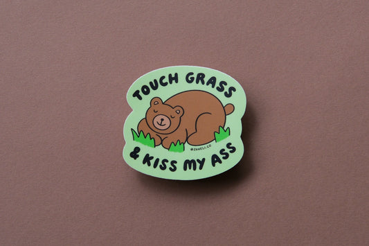 A JaneLi.Co sticker of a napping brown bear that says "Touch grass and kiss my ass" over a brown background.