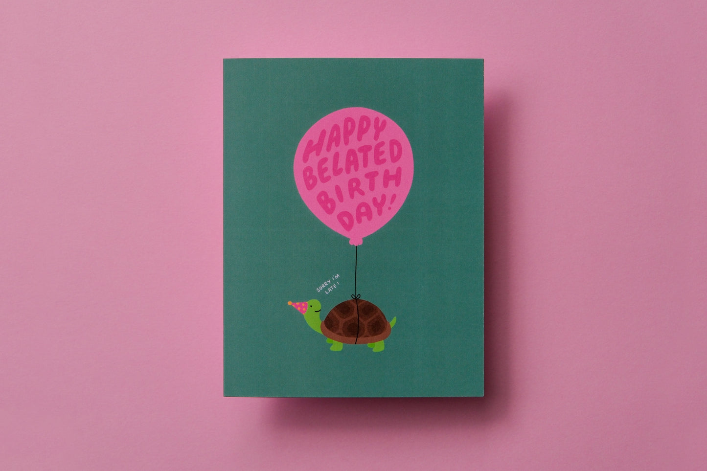A photo of a greeting card with a turtle carrying a balloon that says "Happy belated birthday!" on a pink background.