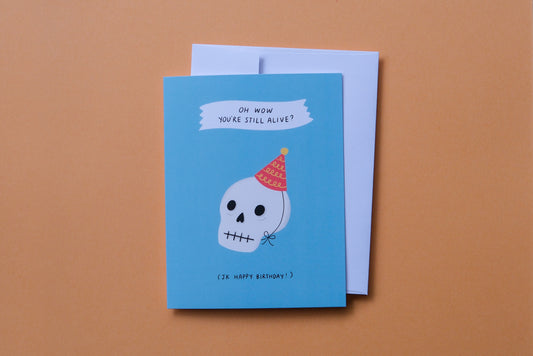 A photo of a greeting card that says "Oh wow you're still alive? Jk happy birthday!" with a skull wearing a birthday hat and a white envelope on an orange background.