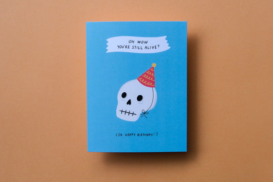 A photo of a greeting card that says "Oh wow you're still alive? Jk happy birthday!" with a skull wearing a birthday hat on an orange background.