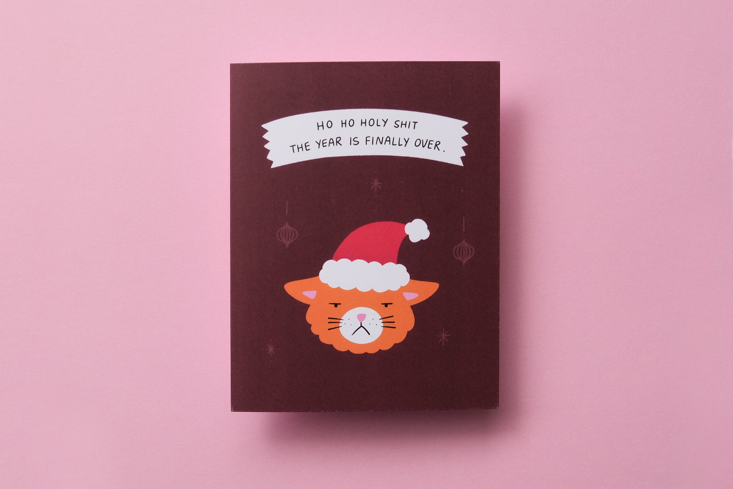 A photo of a greeting card with grumpy santa cat that says "Ho ho holy shit the year is finally over" and on a pink background.