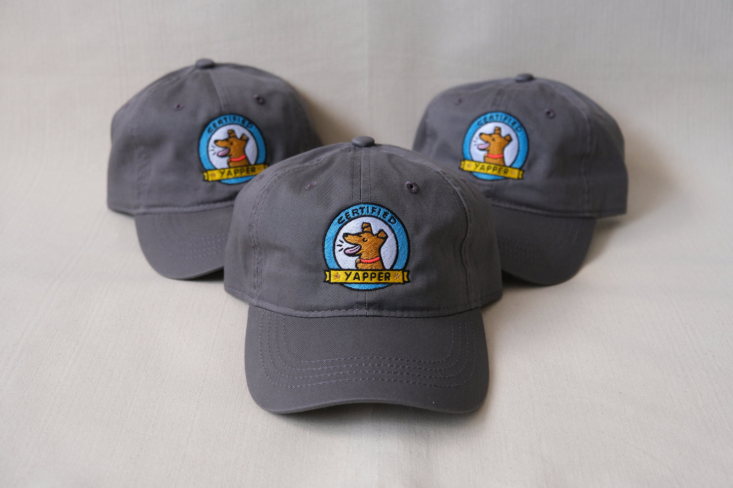 A photo of a 3 grey dad hats embroidered with dogs that say "Certified Yapper" over a cream background.