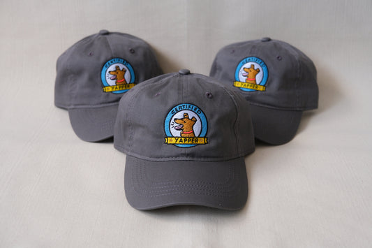 A photo of a 3 grey dad hats embroidered with dogs that say "Certified Yapper" over a cream background.