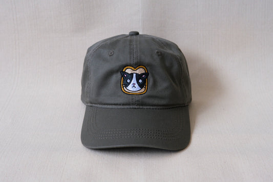 A photo of an olive green dad hat with an embroidered cat stuck in a slice of bread over a cream background.