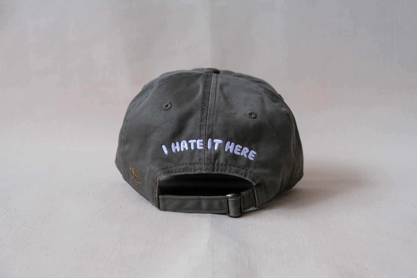 A photo of the back of an olive green dad hat that says "I Hate It Here" embroidered in white with an adjustable buckle strap.