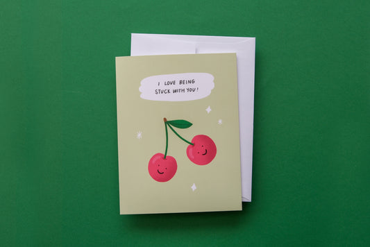 A photo of a greeting card with 2 cherries that says "I love being stuck with you" and a white envelope on a green background.
