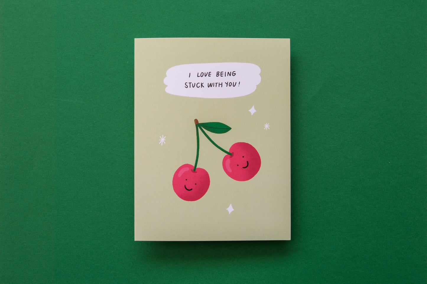 A photo of a greeting card with 2 cherries that says "I love being stuck with you" on a green background.