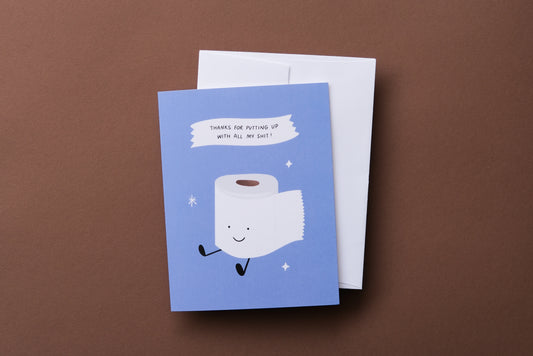 A photo of a greeting card with a smiling roll of toilet paper that says "Thanks for putting up with all my shit!" on a brown background.