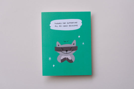 A photo of a greeting card with an evil raccoon that says "Thanks for supporting all my (bad) decisions" and a white envelope on a cream background.