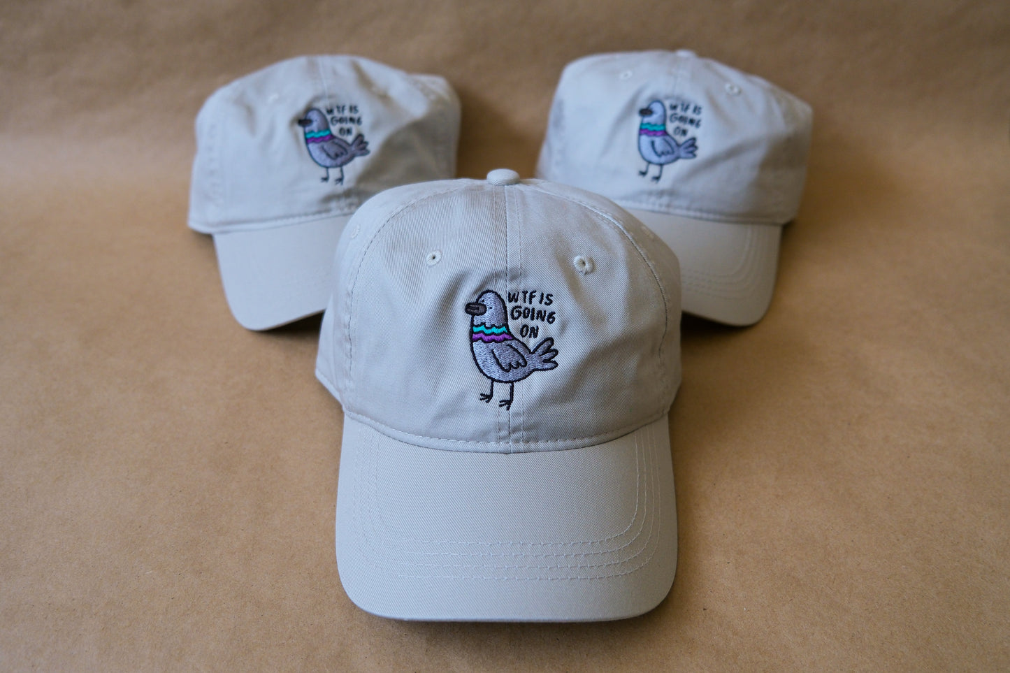 A photo of 3 khaki dad hats with embroidered pigeons that say "WTF Is Going On" over a tan background.