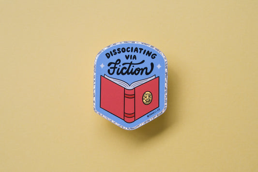 A JaneLi.Co sticker of badge with a book that says "Dissociating via Fiction" over a dark yellow background.