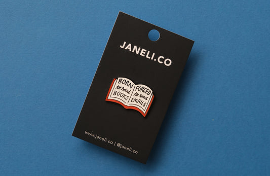 An enamel pin of an open book that says "Born to read books, Forced to read emails" over a blue background.