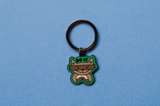 An enamel keychain of an evil raccoon that says "Do it." over a blue background.